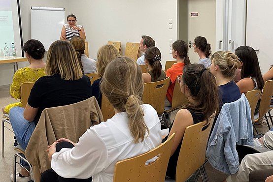 In einem Seminarraum hören viele Zuhörerinnen der Professorin zu.
